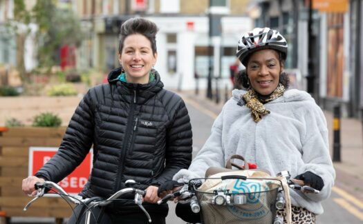 A pair of women smiling with their bikes and looking at the camera. This is one of our Cycle Buddies helping a new cyclist learn a route.